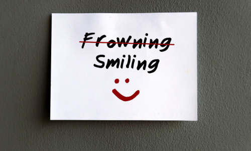 A white paper note with the text frowning and smiling, with frowning struck out in red, and a red smiley face on a grey background