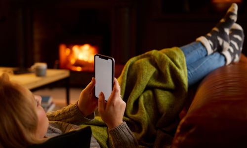 A photo of a woman on a couch in fluffy socks under a blanket with a mobile phone in hand and a fireplace in the background