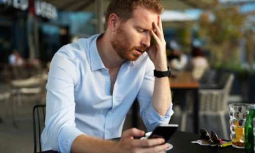 A photo of a stressed out man holding his smartphone sitting at a black table with a blurred out background