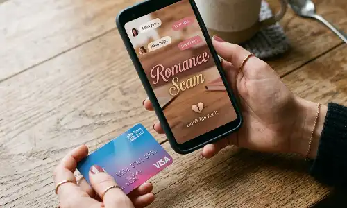 A photograph of hands holding a smartphone displaying "Romance Scam" and a credit card over a wooden background