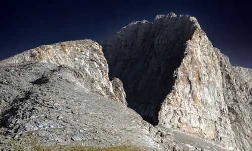 A photograph of the rocky summit of Mount Olympus with jagged ridgelines and distant peaks on a clear blue sky background